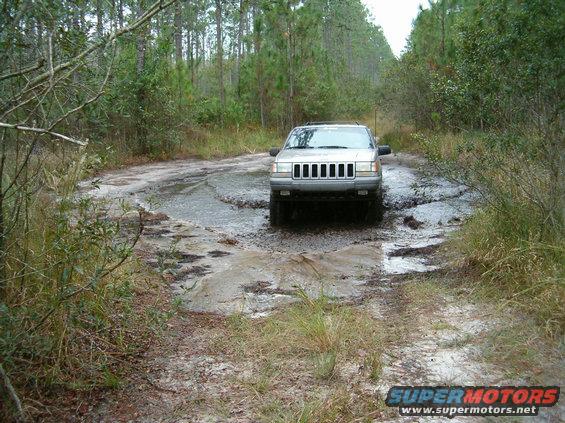 Silver Jeep Grand Cherokee navigating a muddy, pothole-filled dirt road in a wooded area. Water splashes around the tires