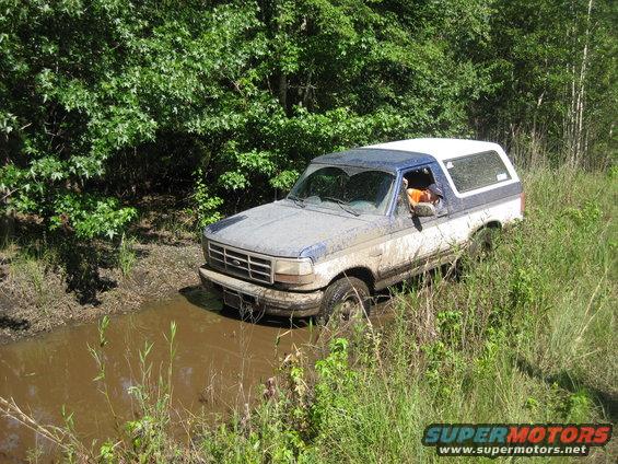 Muddy blue and white Ford Bronco driving through a muddy ditch with trees and tall grass on either side
