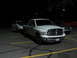 1992 Ford Taurus - A white pickup truck is parked in a lot at night, towing a car on a trailer behind it.