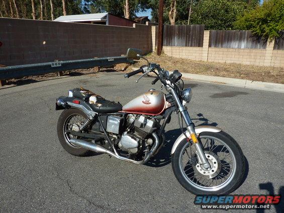1985 Honda CMX250 Rebel motorcycle, red and silver, torn seat, parked on asphalt