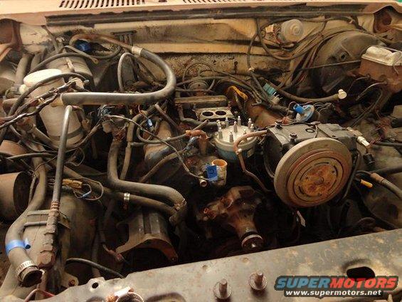 Engine bay with a V8 engine, carburetor, distributor, and air conditioning compressor. Wires and hoses are visible