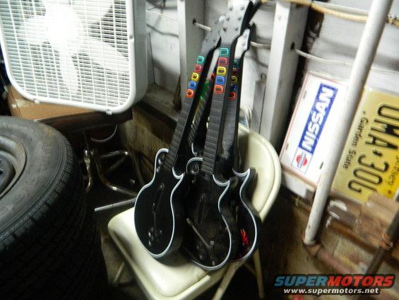 Three black video game guitars with colored buttons are leaning on a chair next to a fan and a spare tire. A Nissan sign and a license plate are on the wall