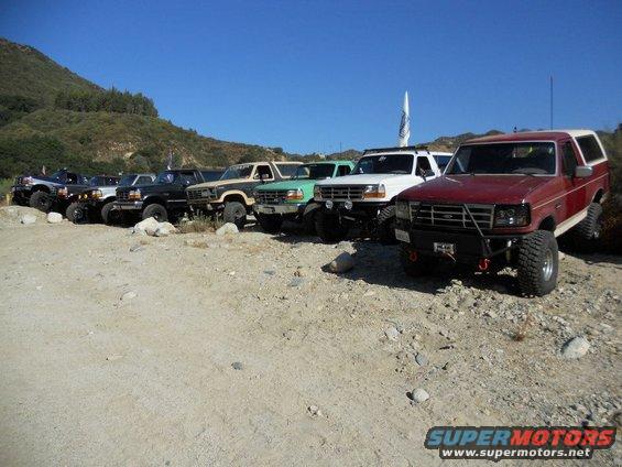 Several Ford Broncos parked on a dirt lot with hills in the background. The red Bronco in front has a custom bumper
