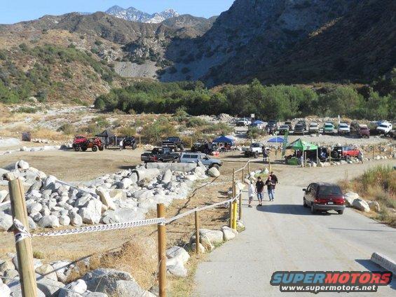Several off-road vehicles parked near tents in a rocky, mountainous area. People are walking on a paved path