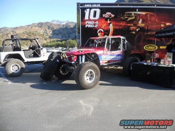 Two custom off-road trucks parked in front of a trailer with racing graphics. One truck is red with BFGoodrich tires, the other is white