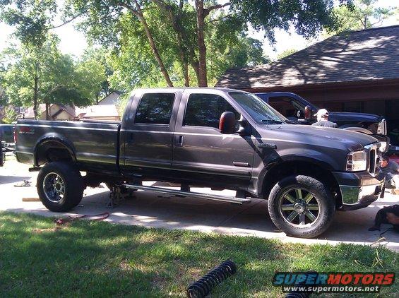 Gray Ford Super Duty pickup truck with aftermarket wheels, jacked up with suspension parts on the ground. Another truck is parked behind it