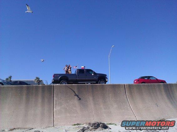 Dark gray Ford F-350 Super Duty pickup truck with two people standing in the bed, Texas flag visible. Red convertible parked to the right