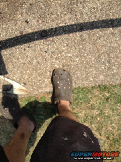 Person wearing dark Crocs and black shorts, casting a shadow on concrete and grass
