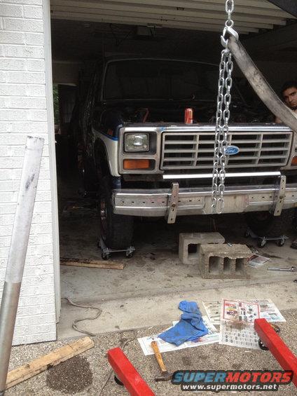 Front view of a dark blue and white Ford Bronco with its hood open, supported by an engine hoist. The vehicle is in a garage