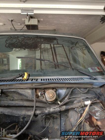 Vehicle windshield with wiper blades and reflection, showing engine bay components below
