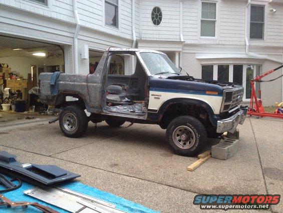 Ford Bronco with doors removed, hood up, and body in rough shape. It's parked in a driveway in front of a house