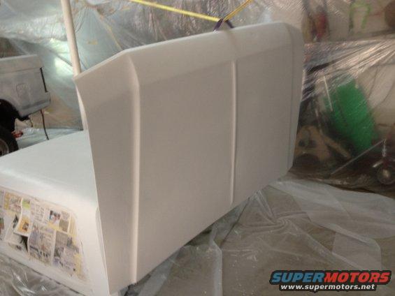 White vehicle hood and fender covered in primer, with another vehicle in the background also being worked on