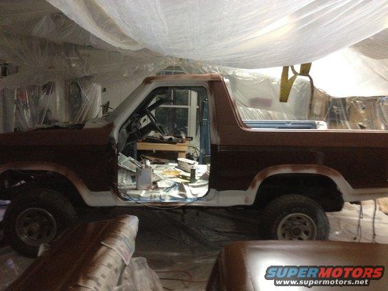 Brown Ford Bronco in a garage, door open, undergoing repairs. White primer visible on body panels