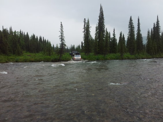 Two SUVs drive through a shallow river with a forest in the background