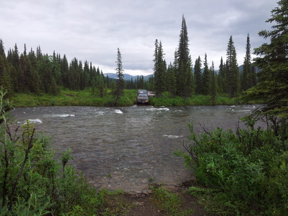 Two SUVs drive through a wide, shallow river surrounded by dense pine forest under a cloudy sky
