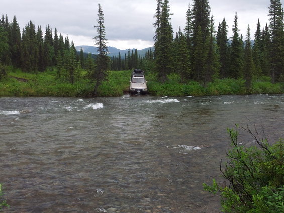 Tan SUV crossing a river with trees and mountains in background