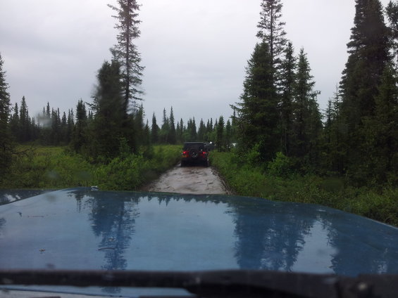 Blue vehicle hood reflects trees and sky, with two SUVs driving on a muddy forest road ahead