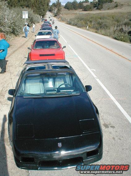 Black 1986 Pontiac Fiero in front of a line of other Fieros parked on the side of a road