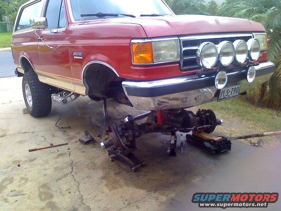 Red Ford Bronco on jack stands with front axle removed, four auxiliary lights mounted on grille guard