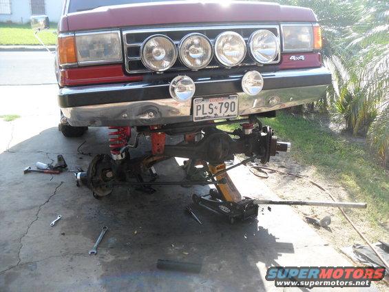 Red Ford Bronco front end with suspension disassembled, sitting on jacks and jack stands. Four auxiliary lights are mounted above the bumper