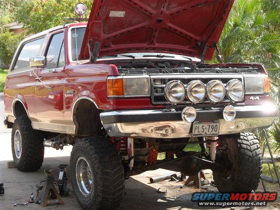 Red lifted SUV, hood open, jacked up with front end work. Has four round lights on the bumper