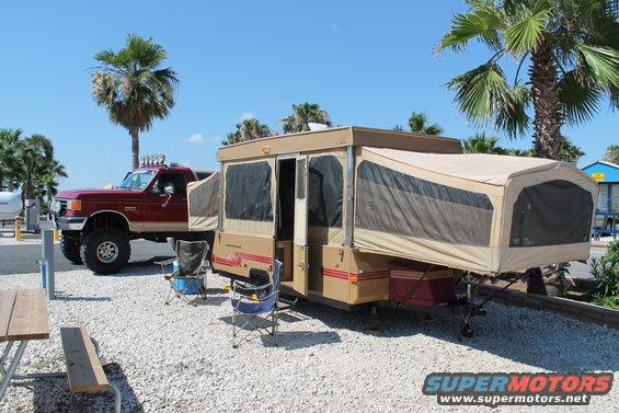 Red 1990 Ford Bronco with lifted suspension and off-road lights parked next to a tan pop-up camper