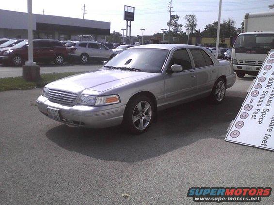 Silver 2003 Ford Crown Victoria parked outside a building with other cars and a truck nearby. Signage is visible on the right