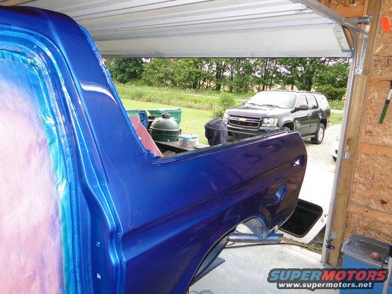 Side view of a blue vehicle body with fresh paint, parked in a garage with another SUV outside