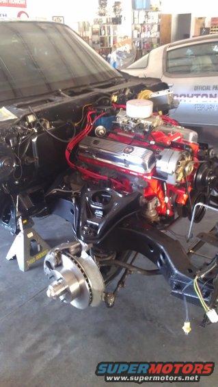 Engine bay with chrome valve covers and red wiring, car on jack stands