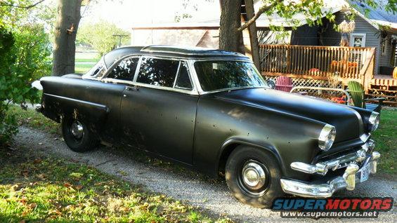 Black 1954 Ford Crestline sedan with matte paint, chrome trim, and visible dents, parked on a gravel driveway