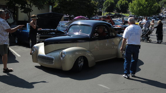 Custom coupe with black roof and tan body, parked outside with people around. Another car with hood up is nearby