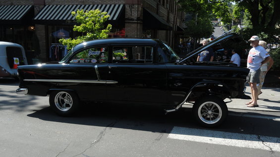 Black 1955 Chevrolet sedan, side view, hood open, chrome trim, custom wheels, people standing nearby