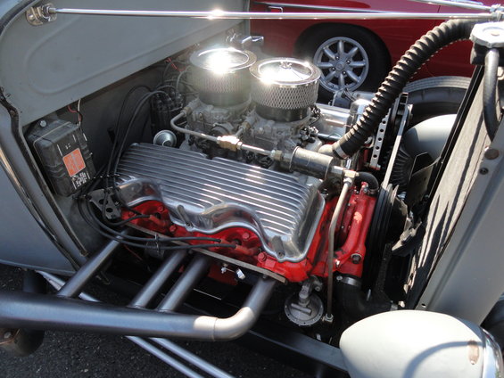 Hot rod V8 engine with dual carburetors, chrome valve covers, and custom exhaust headers. Red engine block visible. Another car's wheel and fender in background