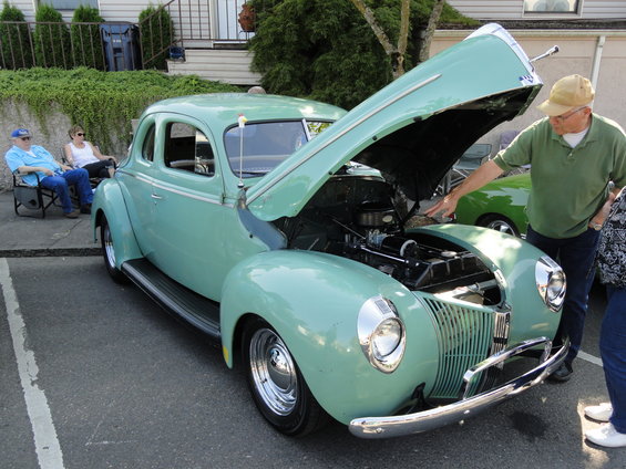 Mint green 1940 Ford coupe with hood open, showing engine. Man in green shirt points to engine. Another car visible behind