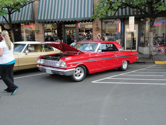 Red 1964 Ford Fairlane with hood open parked next to a tan car. Street scene with shops in background