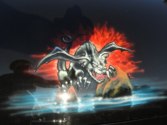 Black car hood with airbrushed mural of a grey dragon breathing fire, with a silhouette of a person on the left