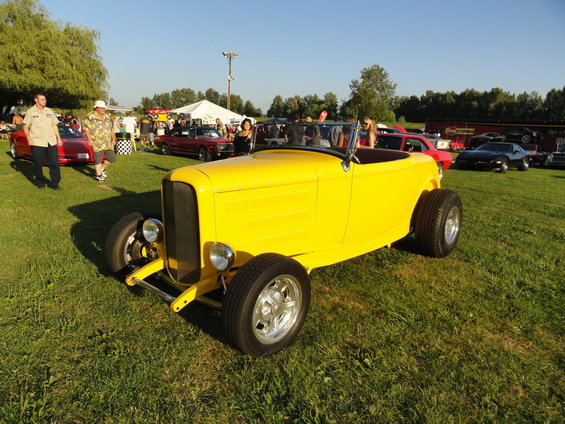 Yellow hot rod convertible with wide rear tires parked on grass at an outdoor car show. Other cars and people are visible in the background