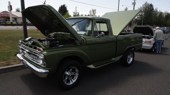 Green 1965 Ford F-series pickup truck with hood and bed open, parked next to a white car