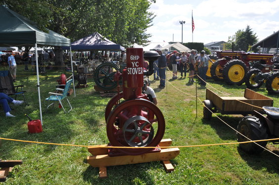 Red stationary engine with text '6HP YC STOVER' on a wooden base, with tractors and tents in the background