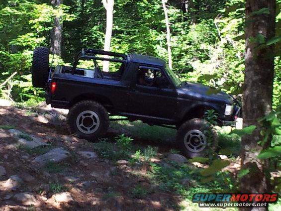 Black SUV with external spare tire driving on a rocky, wooded trail. Appears to be a 1990s model