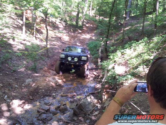 Black off-road SUV driving through a muddy, wooded trail with large tires and extra lights on the front