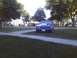 2003 Ford Crown Victoria - A blue Ford Mustang is parked on the grass near a gathering of people under trees.