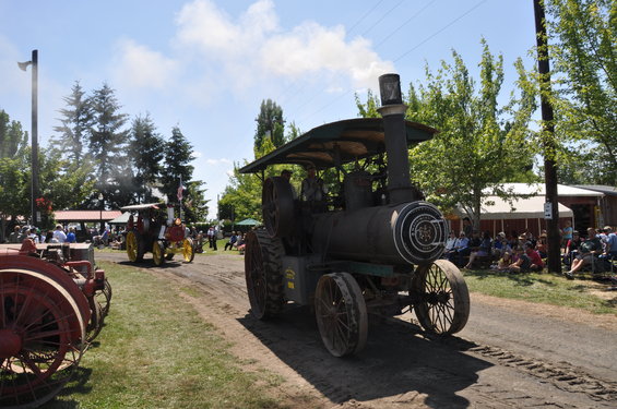 Steam tractor with canopy and large wheels, emitting smoke, driving on a dirt road past spectators