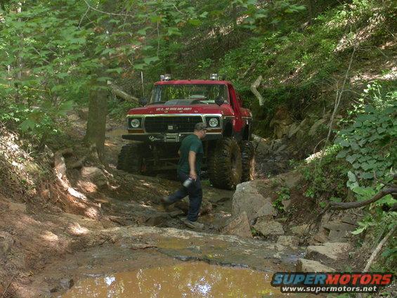Red 1979 Ford F-Series truck with large off-road tires navigating a muddy, rocky trail. A person walks beside it