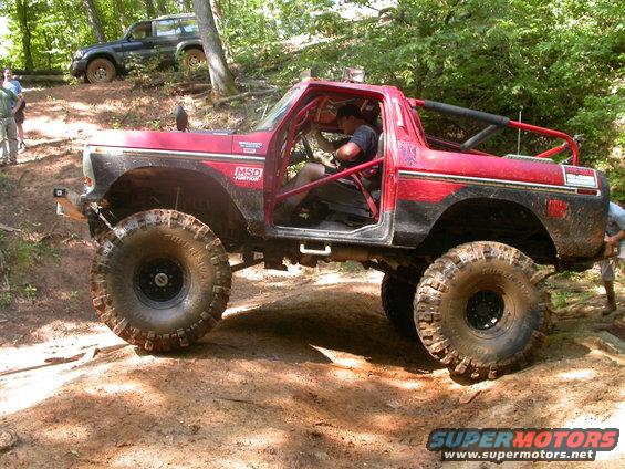 Red 1990 Ford Bronco with roll cage and large tires on a dirt trail