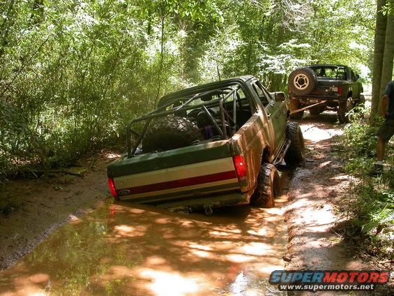 Two off-road vehicles stuck in a muddy trail in the woods. One is green with a roll cage, the other is black and being towed