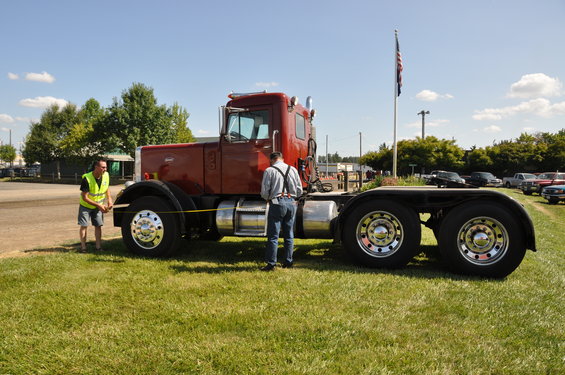 Man in yellow vest measures front wheel of red Peterbilt 359 truck. Another man stands by the cab