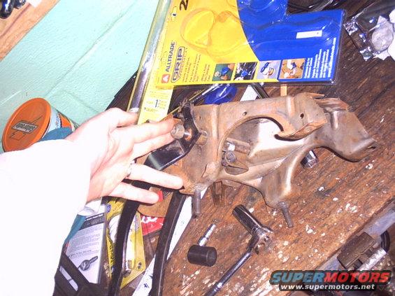 Close-up of a rusty car pedal assembly with a hand pointing to a bolt. Tools and packaging are on a wooden surface
