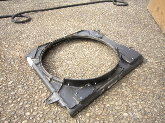 Black plastic radiator fan shroud with mounting tabs and screw-on covers, lying on a gravel surface