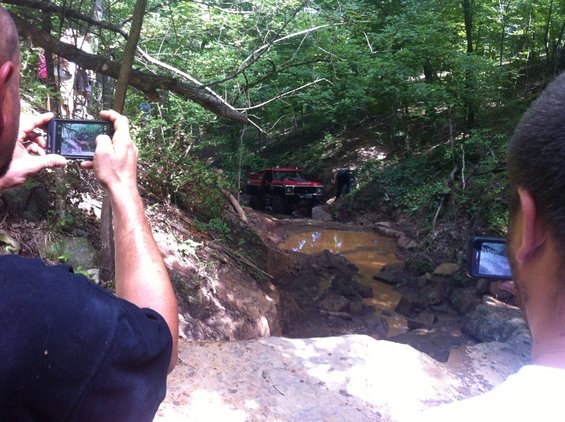 Red off-road truck driving through mud pit on wooded trail. Two people filming with phones